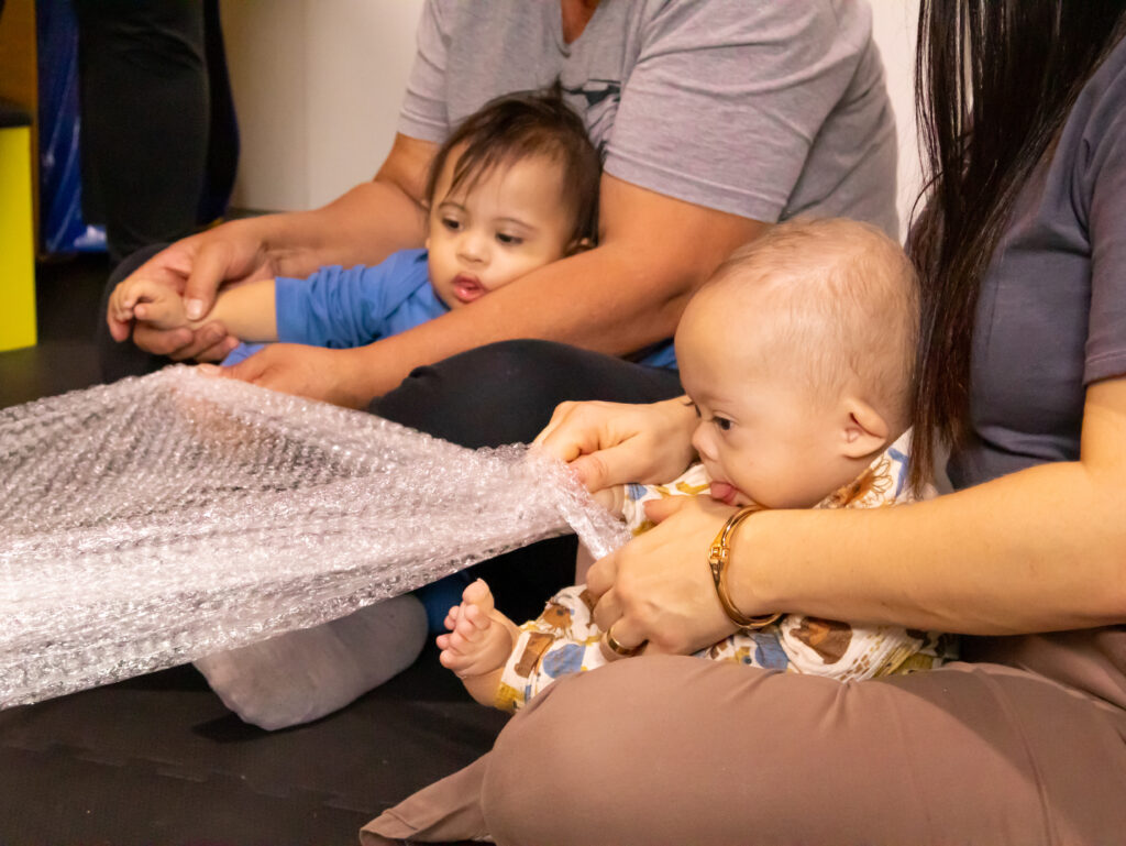 Duas crianças pequenas participam de uma atividade sensorial, acompanhadas por adultos. Uma das crianças veste roupa azul e está sentada no colo de uma pessoa que segura sua mão. A outra criança, de roupa estampada, está no colo de uma mulher e segura uma folha de plástico bolha com curiosidade, aproximando o rosto para explorá-la com a boca. A cena transmite cuidado, estímulo e interação afetiva.